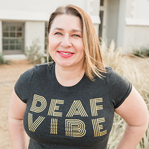 Erin, owner of Mabely Q and a Deafblind entrepreneur smiles into the camera wearing a t-shirt that says 'Deaf Vibe'.