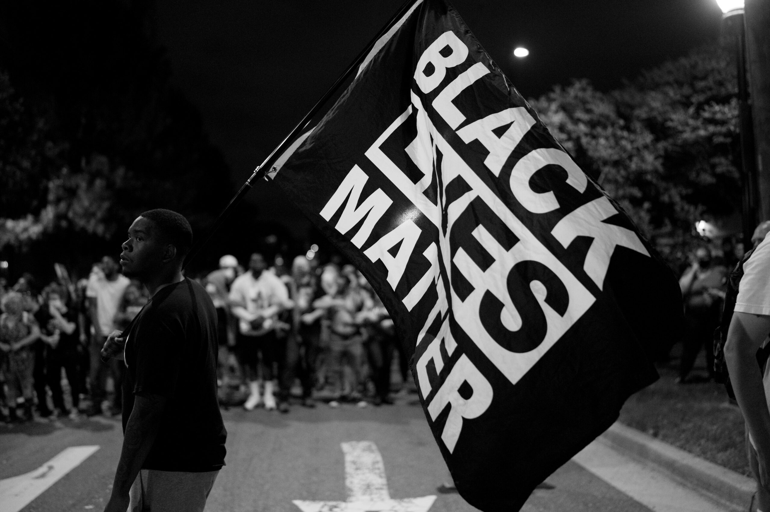 Black Lives Matter protest at night with a person holding a large flag in the street, surrounded by a crowd of protesters.